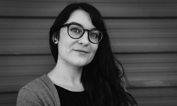 Black and white portrait of Elyse smiling with her back against a textured metal wall.